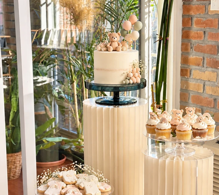 Cake with a teddy bear topper, cupcakes and decorated cookies on cream pedestal stands by a sunny window